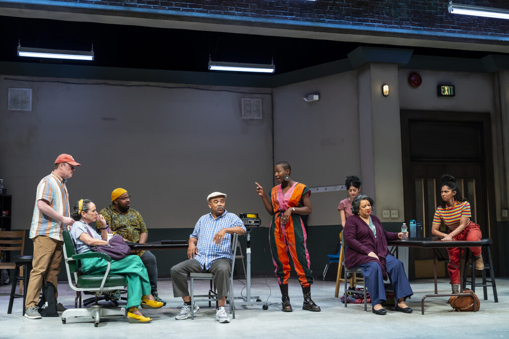 A group of multicultural and intergenerational actors sit and stand at tables in a rehearsal room. One Black actor wearing a brightly-colored jumpsuit stands at center guiding the group, pointing with her right finger.