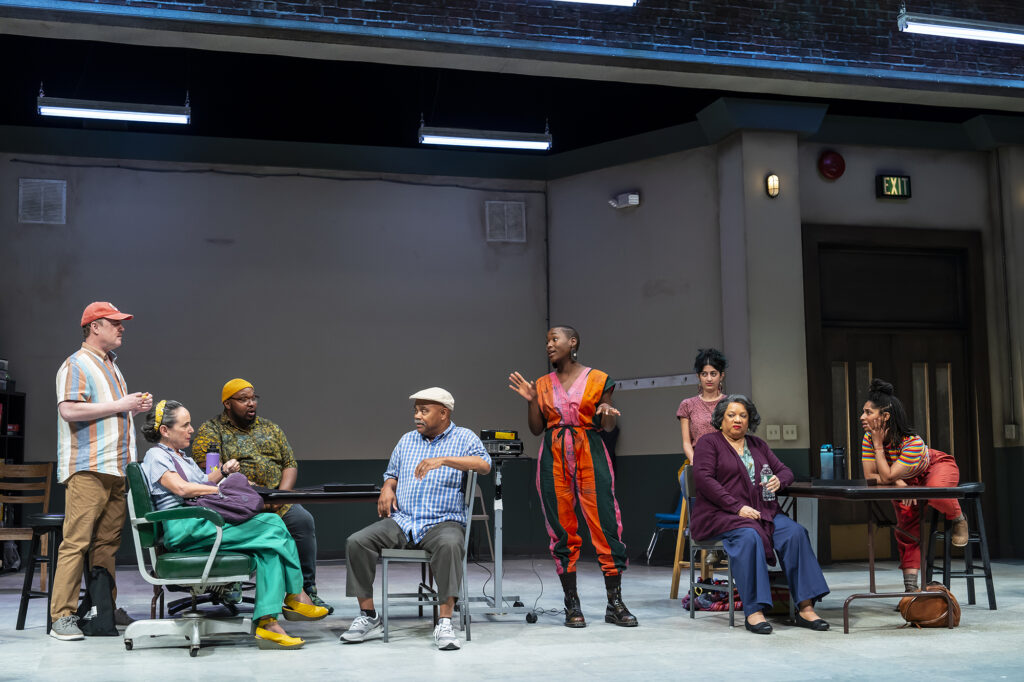 A group of multicultural and intergenerational actors sit and stand at tables in a rehearsal room. One Black actor wearing a brightly-colored jumpsuit stands at center guiding the group, with the palm of her hand raised.