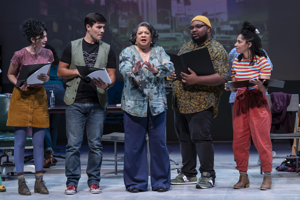 Five multicultural and intergenerational actors stand with binders in hand in a rehearsal room, excitedly retelling the story of the era leading up to Maynard Jackson’s election.