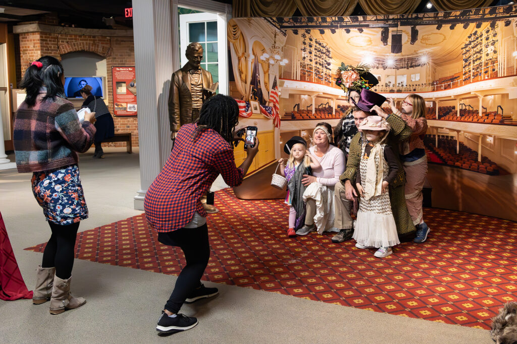 A family with young neurodiverse children dresses up in historical costumes as a Ford’s Theatre staff member takes their photo, in front of a backdrop displaying Ford’s Theatre.