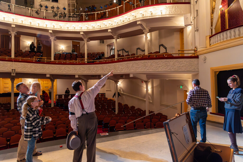 An actor teaches neurodiverse children about Ford’s Theatre, pointing towards the stage right box. Two parents stand to their right.