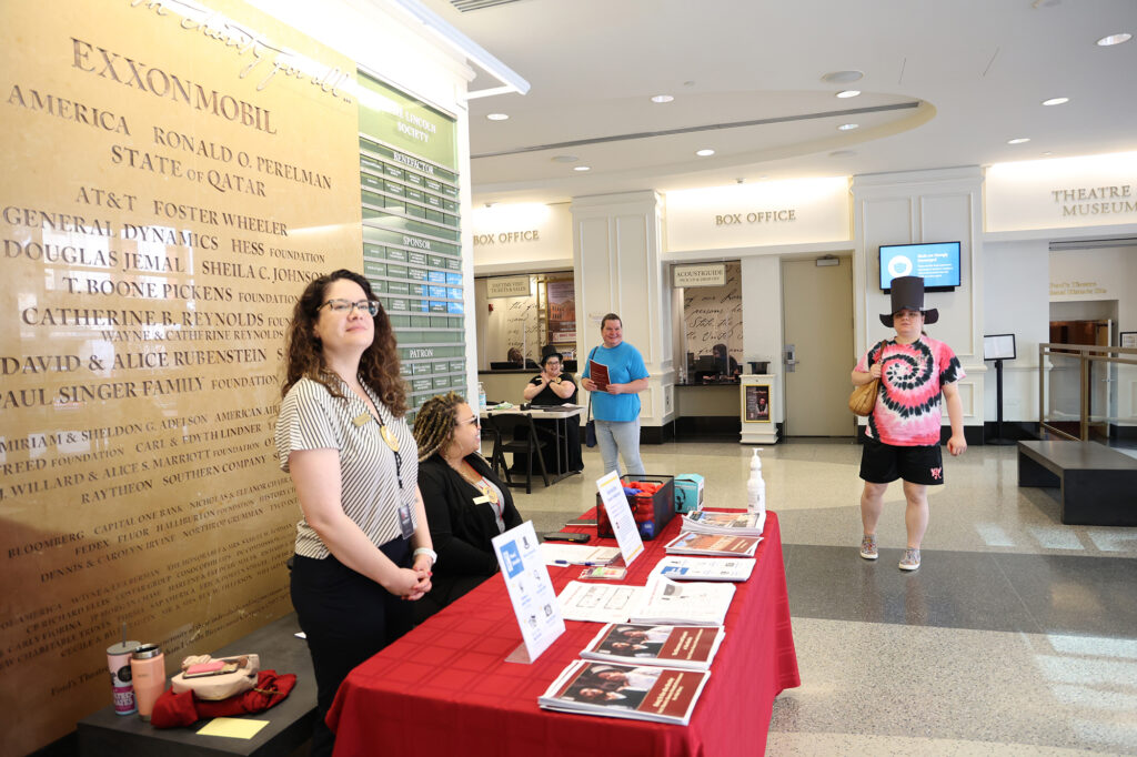 Two women sit at a check-in table, a woman wearing an Abraham Lincoln top hat stands near them with another visitor carrying pre-visit materials, and a staff member also wearing a Lincoln hat sits at a table further away.