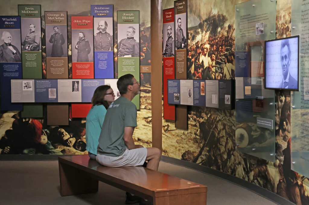 A man and a young girl, both wearing glasses, sit and watch a video in a museum.