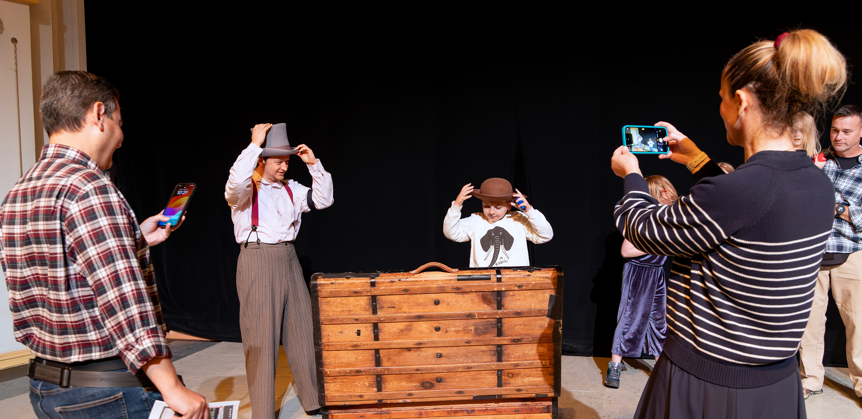 A neurodivergent child and a neurotypical actor put on hats, along with a neurotypical actor, behind an open wooden box on the Ford’s Theatre stage. His parents take photos of them.