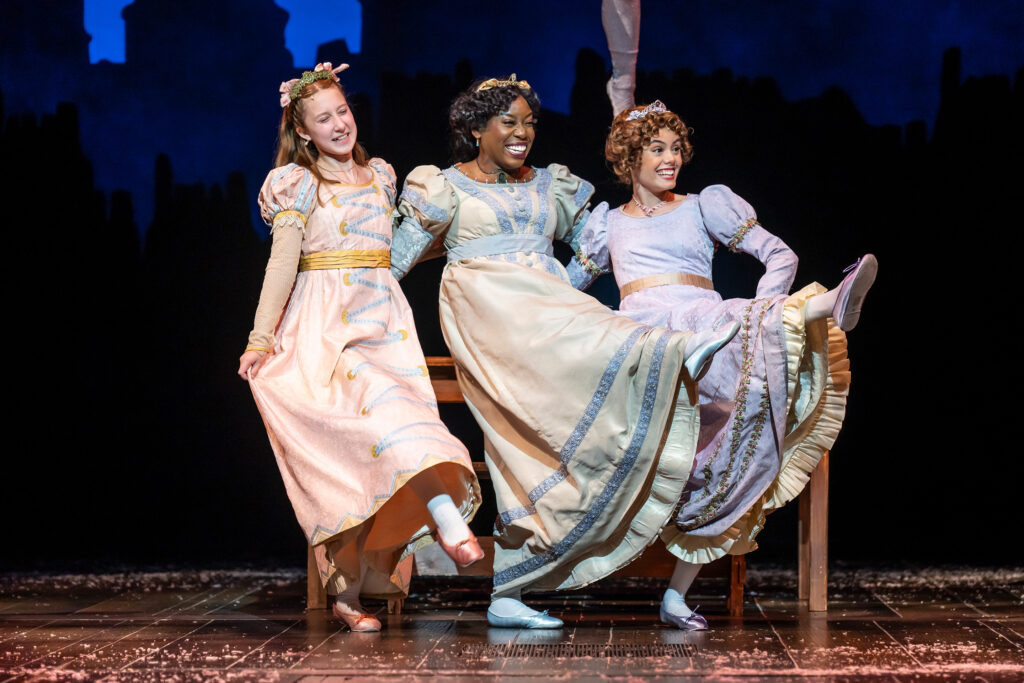 A diverse, multigenerational group of girls wearing Victorian-era dresses smile and dance joyfully, with their left legs kicked up high like a “Can-Can.”