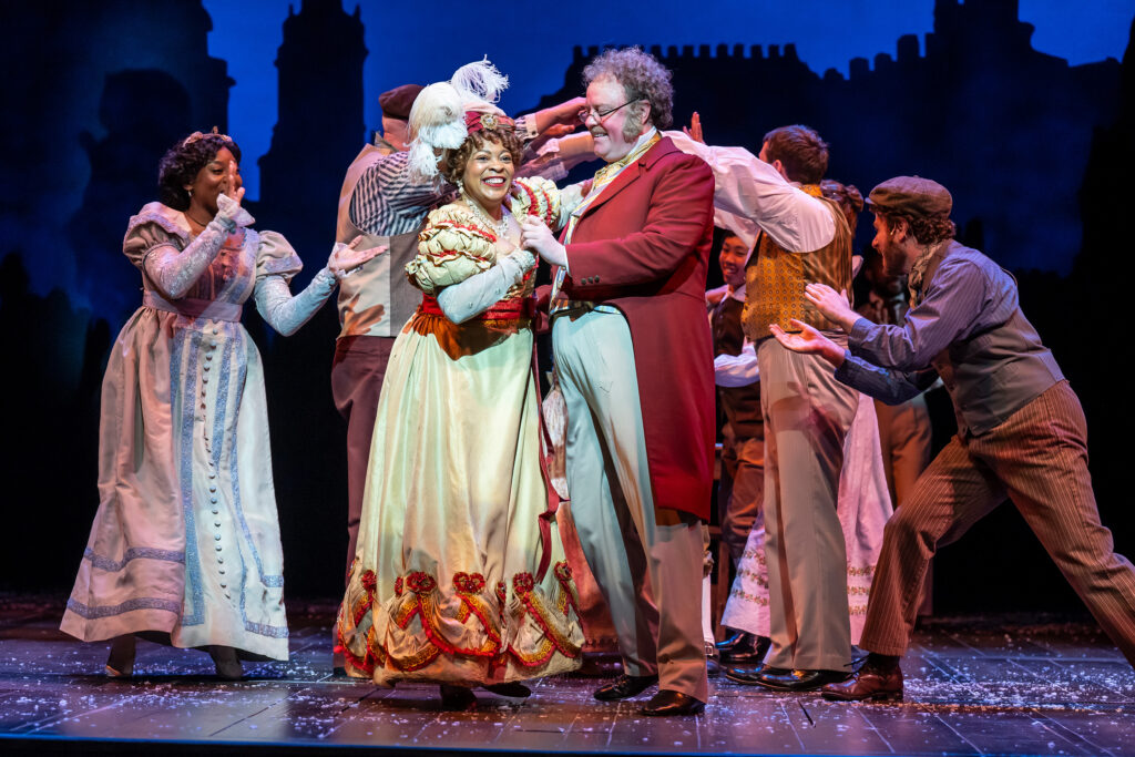 A diverse cast in festive garb dances onstage, with a happy couple holding hands and embracing at center stage.