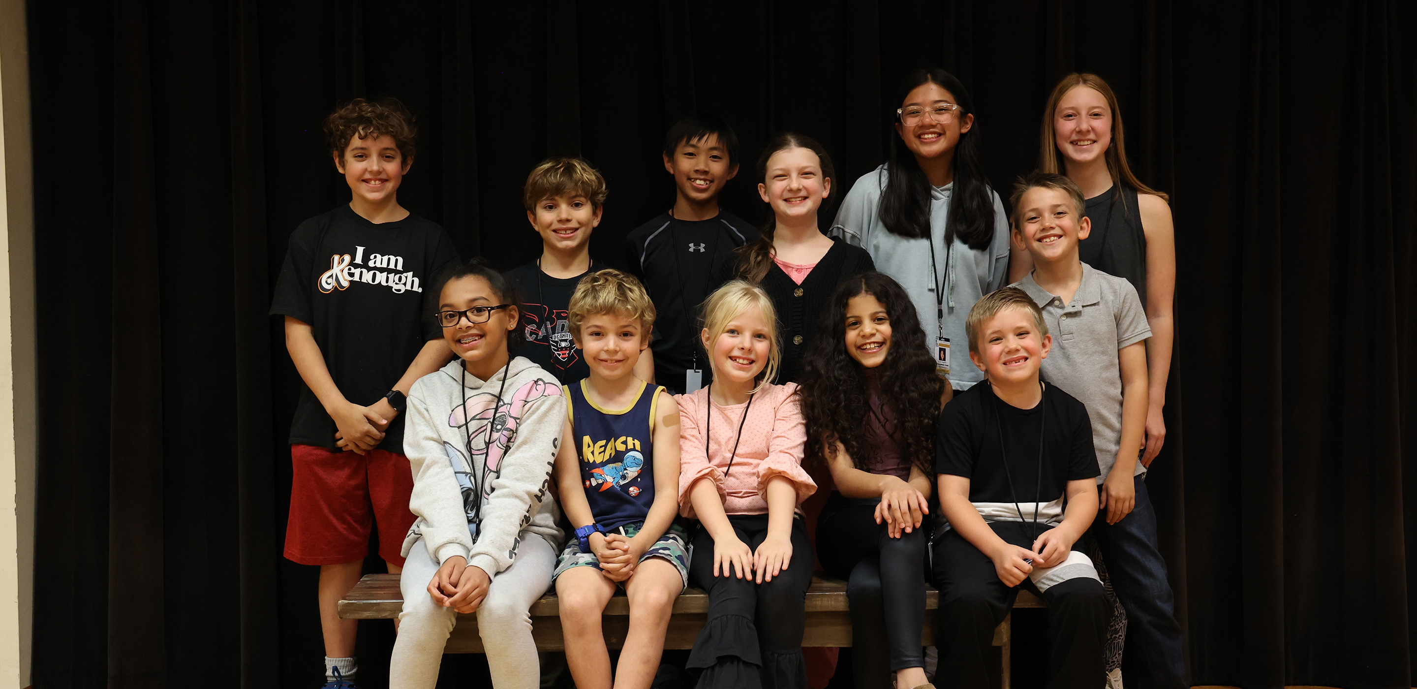 A group of diverse child actors smile excitedly and pose for a group photo at a first rehearsal. Half of the group is standing, and half is sitting on a bench, in front of a black curtain.