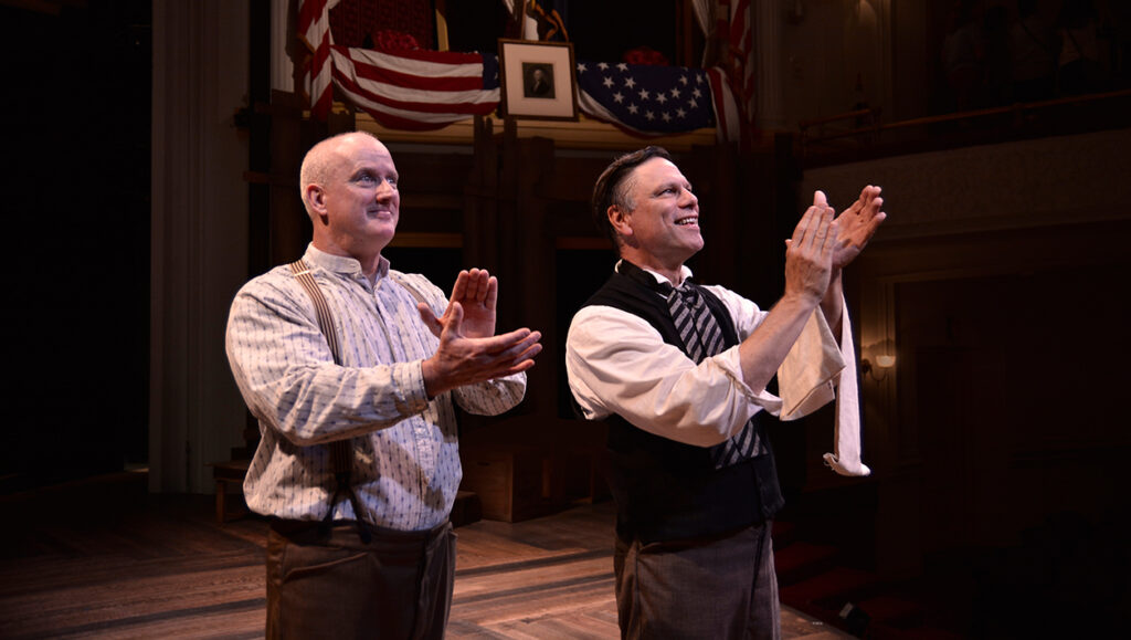 Actors playing Harry Hawk and Harry Ford wear 1860s costumes and mourning arm bands. They stand smiling and clap at someone not pictured in the distance. Behind them is the flag-draped Presidential Box at Ford’s Theatre.