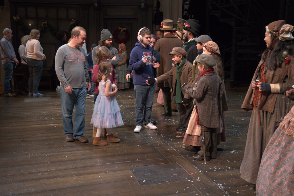 A young boy extends his hand to a young girl, as cast members dressed in Victorian-era clothing greet patrons in a meet and greet onstage.