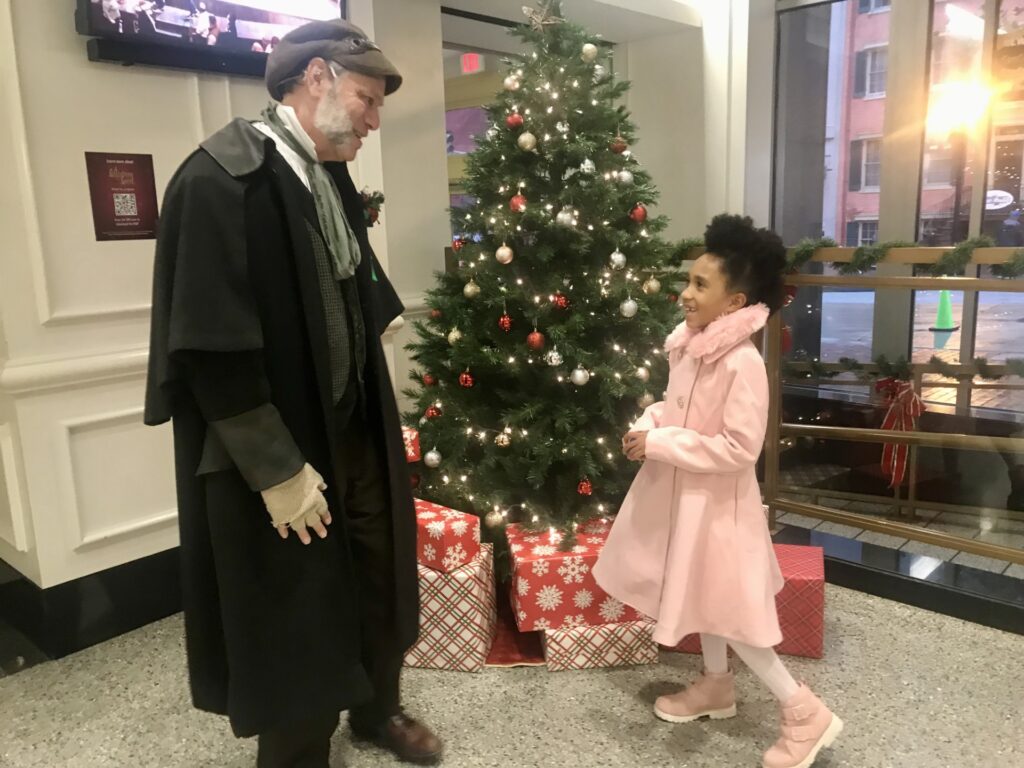 A man dressed in Victorian-era clothing welcomes a young girl in a pink coat to Ford’s Theatre. They stand in front of a decorated Christmas tree.