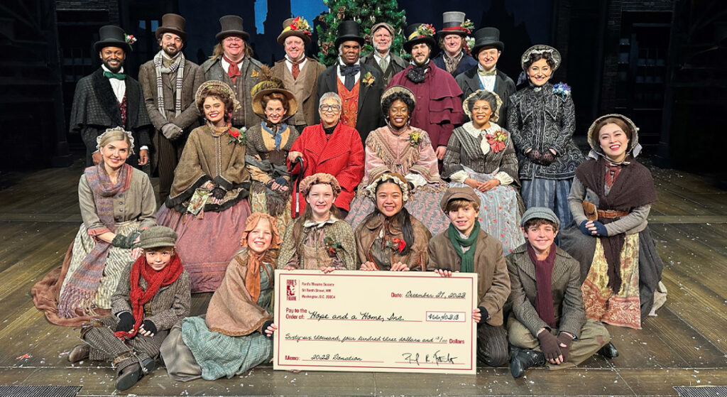 A multigenerational, diverse group of actors wearing Victorian-era clothing and a woman wearing a red coat pose on a stage in front of a Christmas tree, with a large check in the amount of $66,403.18.