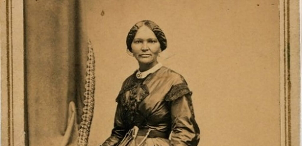 A sepia photograph of an African-American woman in a fancy 19th century dress standing next to a chair over which a curtain has been draped.