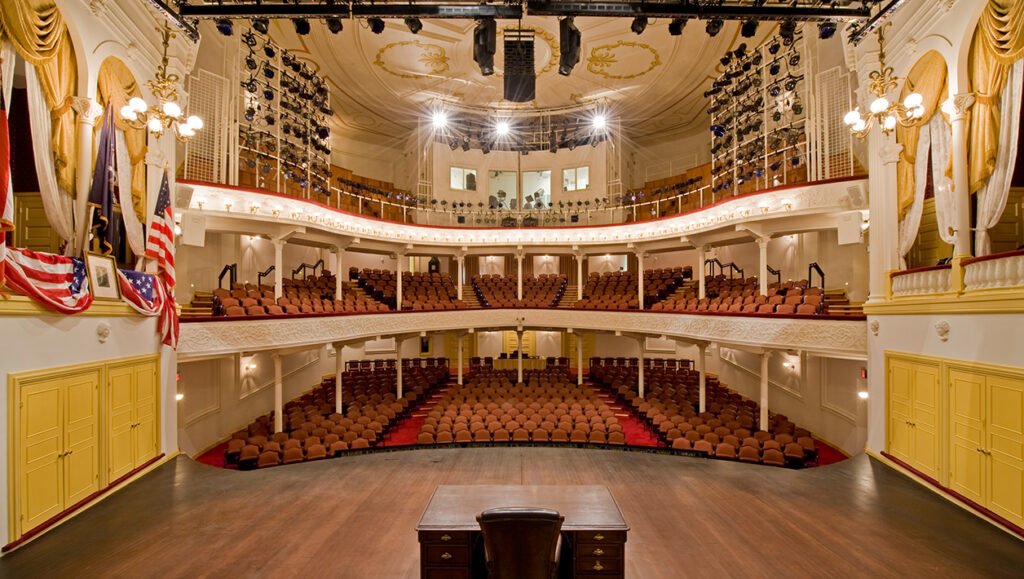 The view from the Ford’s Theatre stage looking out to the audience. To the left of the stage is the President Box with an American flag, a framed picture of George Washington and American flag bunting draped over the box. To the right is another box with yellow and white curtains. In the center of the stage is a wooden desk. The view includes two levels of seating and rows of lighting equipment on the third level.