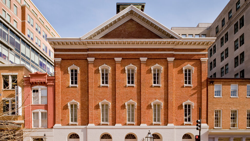An exterior view of historic Ford’s Theatre, a three-story brick building with five arched windows on the second and third floor. The bottom floor is white and has five arched entryways.