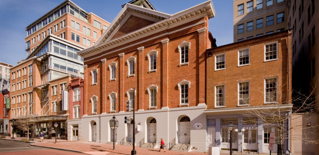 A side exterior view of historic Ford’s Theatre, a three-story brick building with five arched windows on the second and third floor. The bottom floor is white and has five arched entryways.