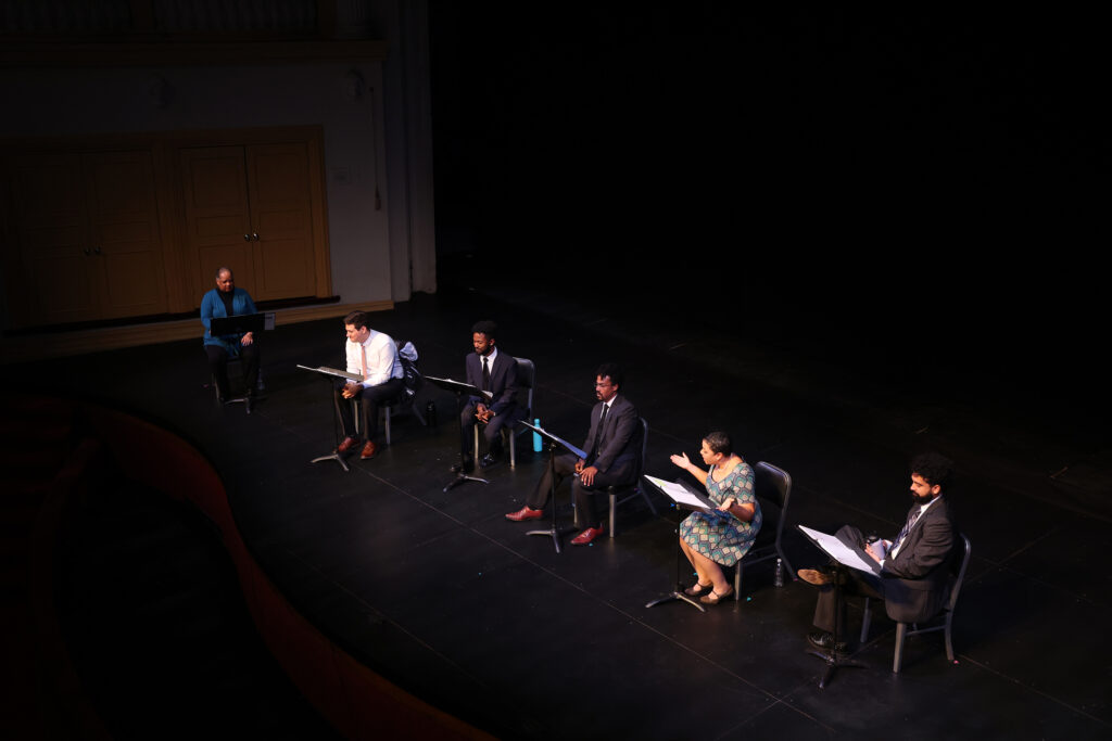 Six diverse actors sit in chairs with music stands and scripts and read a play.
