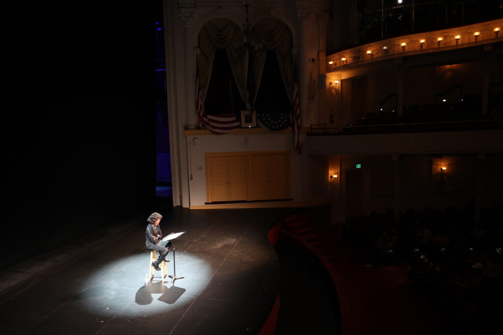 A Black woman sits in a chair on the Ford’s Theatre stage reading her play, in a wide shot that includes the Presidential Box.