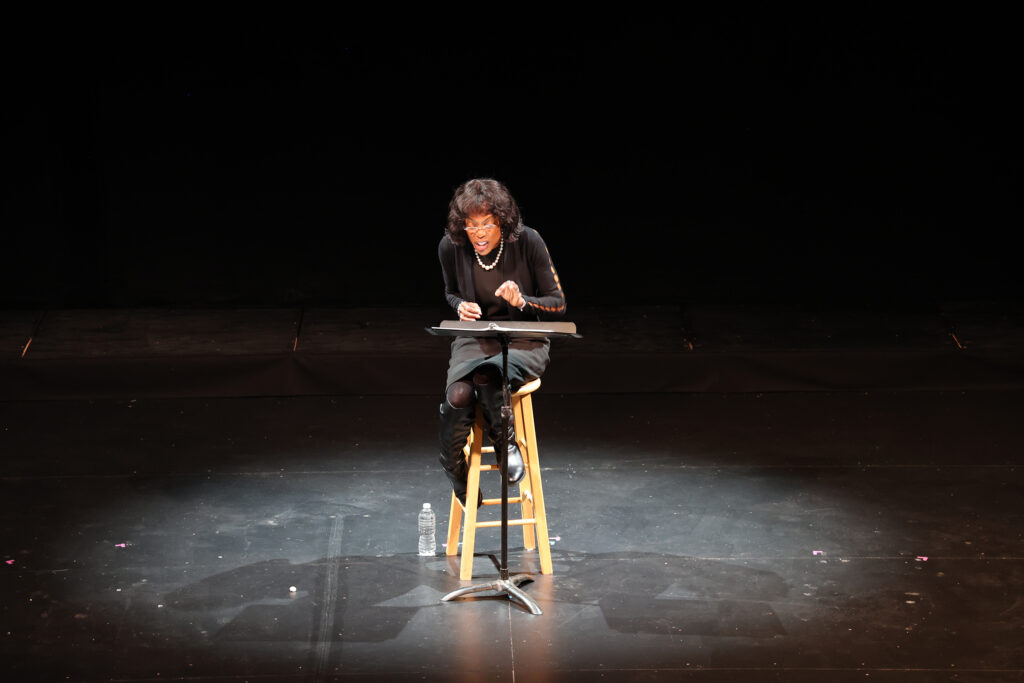 A Black woman sits in a chair with a music stand under a spotlight on the Ford’s Theatre stage, reading her play with an expression of frustration.