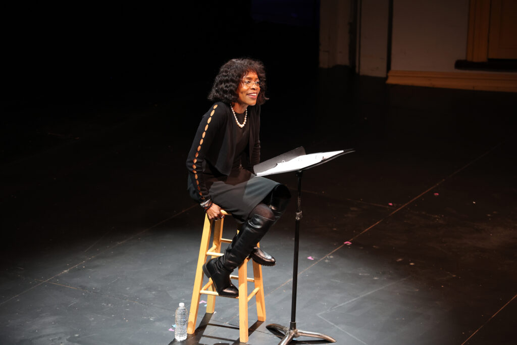 Designer of Note 3 Charlayne Woodard in her 2024 Ford’s Theatre reading of A Designer of Note, A Woman of Style, directed by Delicia Turner Sonnenberg, with dramaturgy by Sydné Mahone. Photo by Rachel Flannery. A Black woman sits in a chair with a music stand under a spotlight on the Ford’s Theatre stage, reading her play and smiling softly.