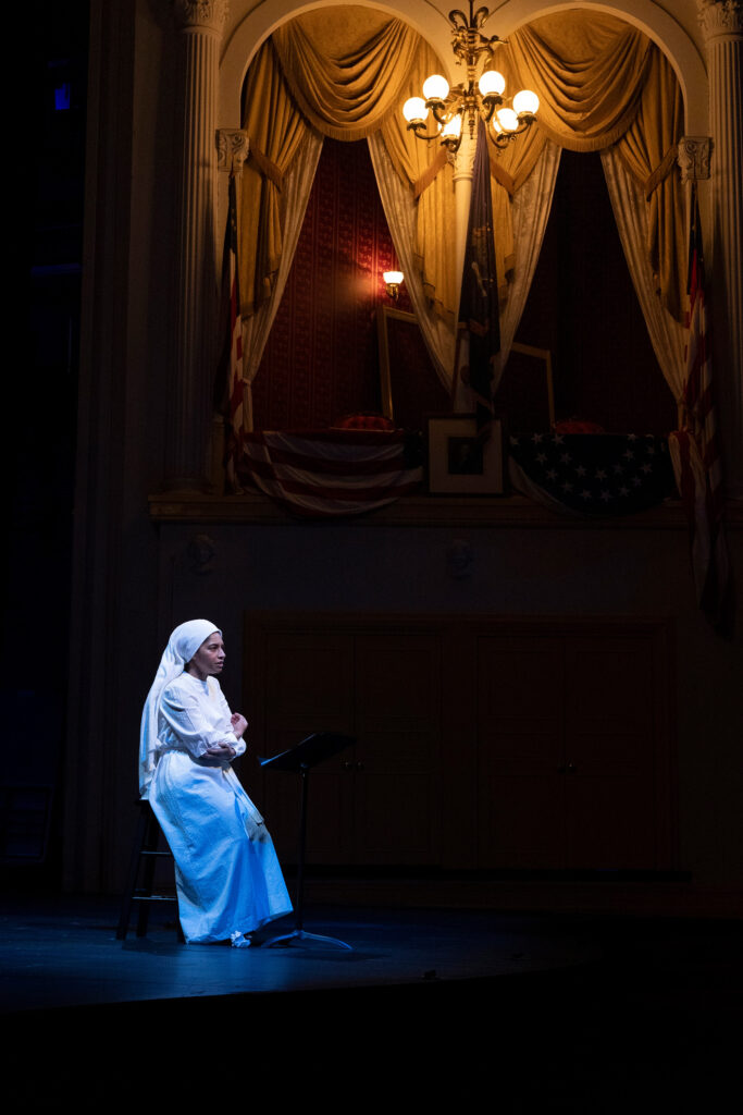 SISTER X 1 Constance Swain as Ameilia in the 2024 Ford’s Theatre reading of SISTER X by Nambi E. Kelley, directed by Hana S. Sharif, with dramaturgy by Sydné Mahone. Photo by Carolina Dulcey. A Black woman sits under blue light. She wears a white head covering and traditional dress of the Nation of Islam. The Presidential Box at Ford’s Theatre is visible in the background.
