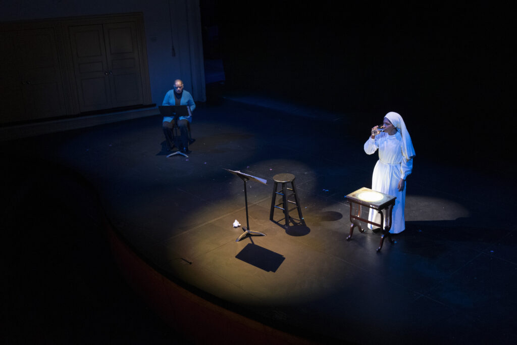 A Black woman drinks a glass of water. She wears a white head covering and traditional dress of the Nation of Islam. A spotlight remains on an empty chair and music stand with a script. Another Black woman sits at the side reading stage directions.
