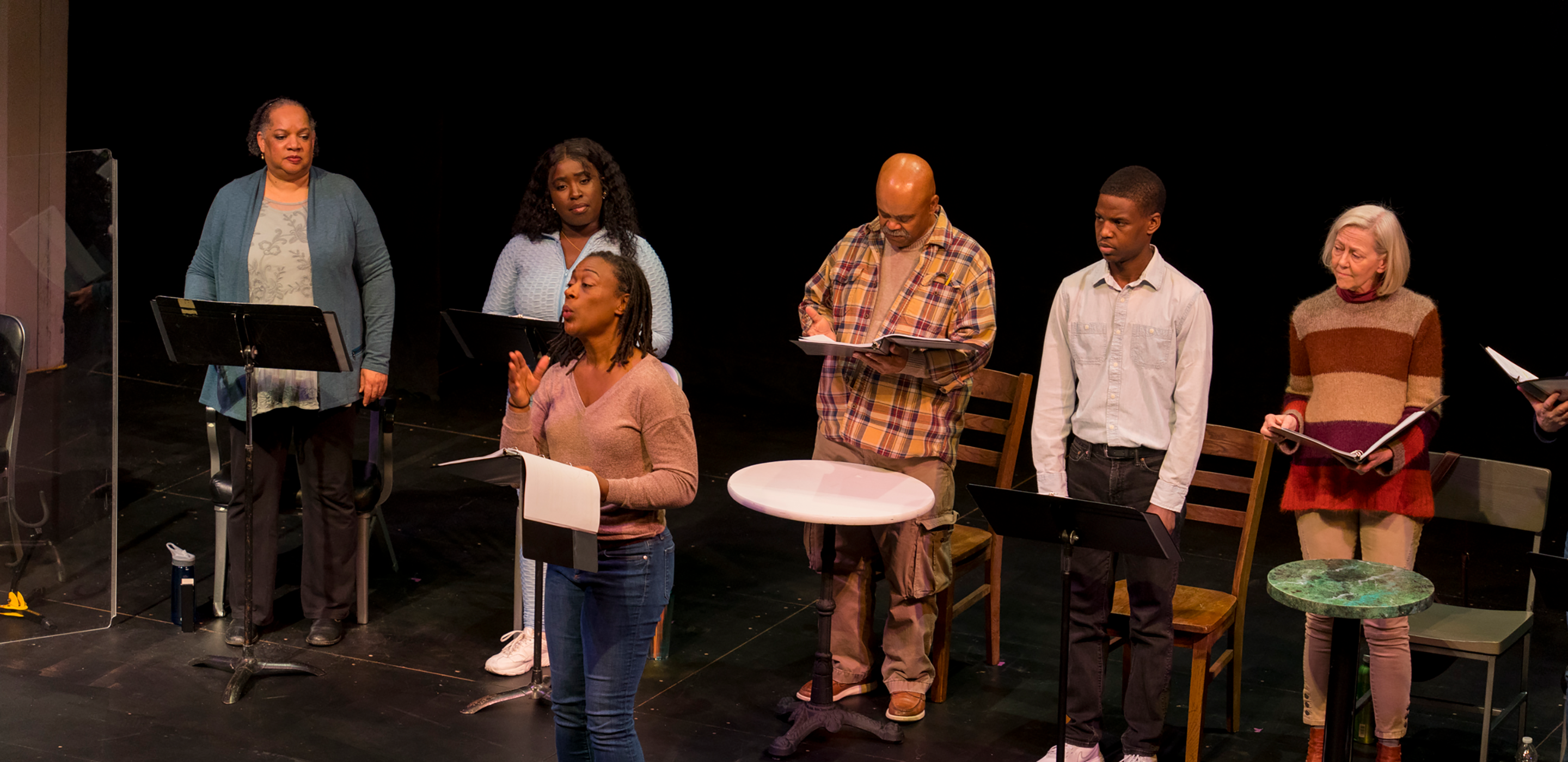 Five actors stand in a line on a stage. In front of them, music stands and tables. Some of them hold binders. At the front of the stage, a woman with short locs wearing blue jeans and a tan sweater holds a binder and speaks to the audience.