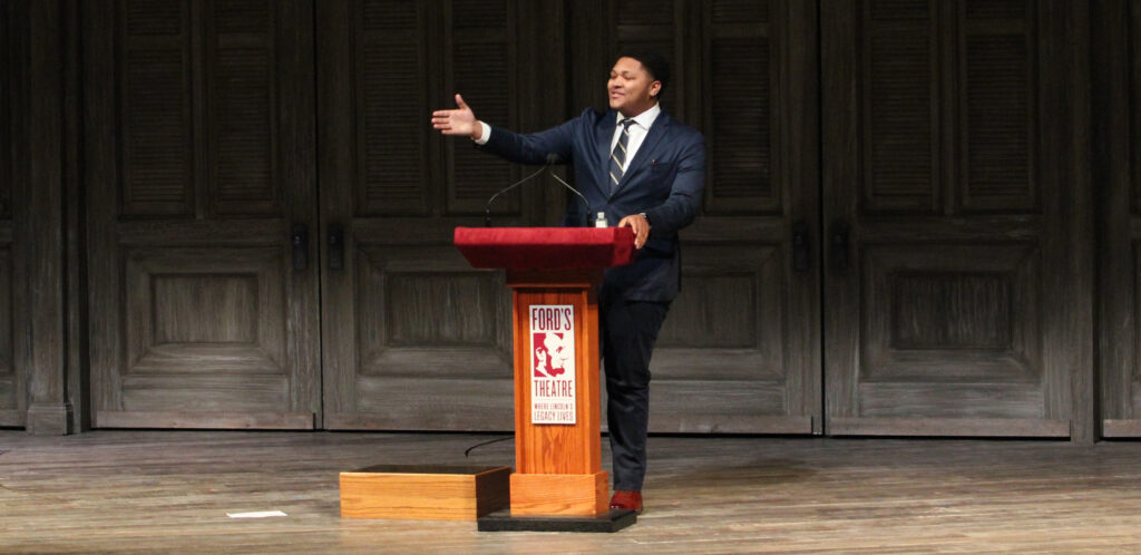 A Black teenager dressed in a suit and tie stands a podium on a stage and gives a speech.