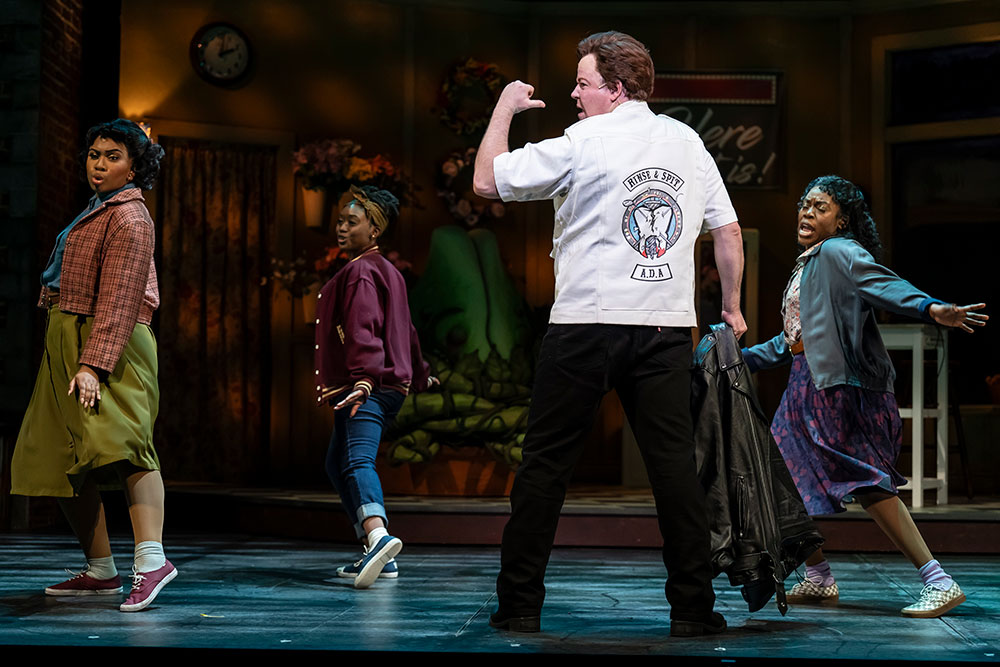 A white man turns his back to an audience and faces sideways, wearing a white dentist shirt that reads “Rinse & Spit A.D.A.” from the back with an image of a tooth. Three Black women in colorful streetwear dance next to him.