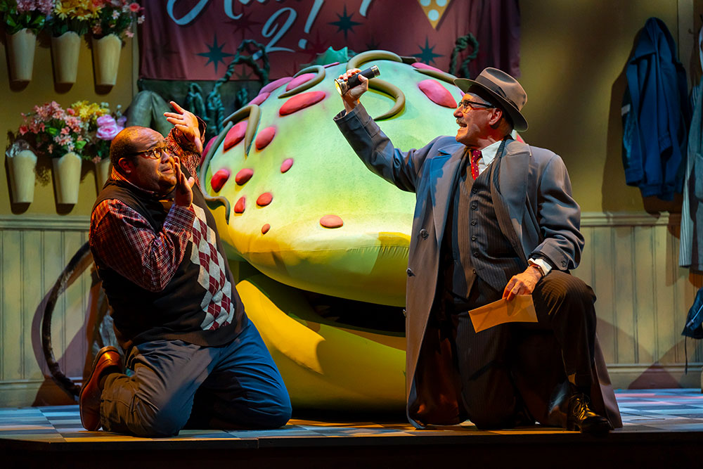 Two men kneel on the floor in front of a large, green strange and interesting plant. One is holding a flashlight and envelope, while the other has an expression of fear.