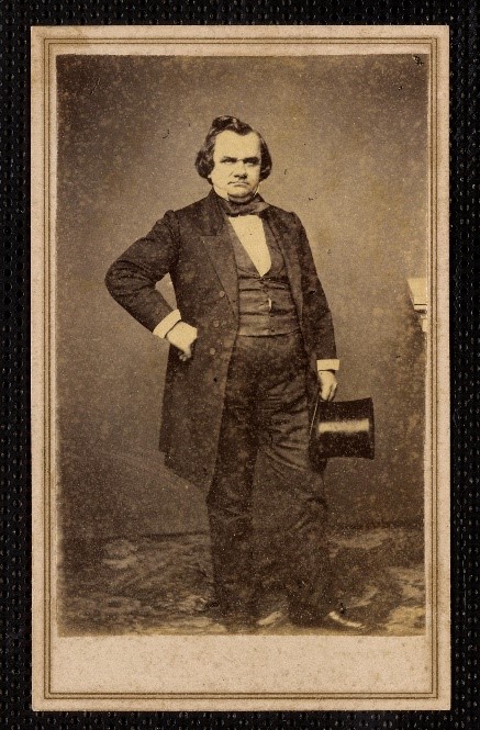 A sepia photograph of a man standing with his right hand on his hip. He is carrying a top hat.