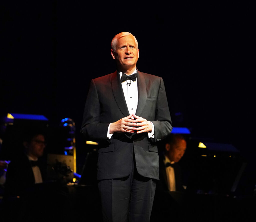 A man in a suit stands and gives a speech in front of a band.
