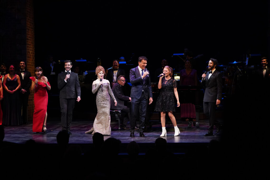 Three men and three woman sing on a stage in formalwear. Behind them a band accompanies.