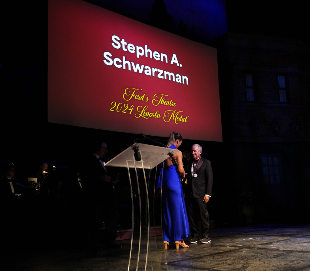 A young woman in a blue dress hands a medal to an older man in a suit. A screen behind them reads, "Stephen A. Schwarzman, Ford's Theatre 2024 Lincoln Medal."