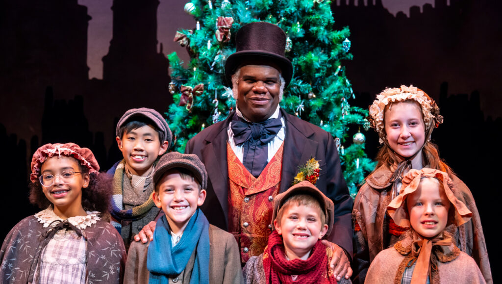 A smiling Black man in Victorian clothing stands in front of a Christmas tree, surrounded by smiling young children.