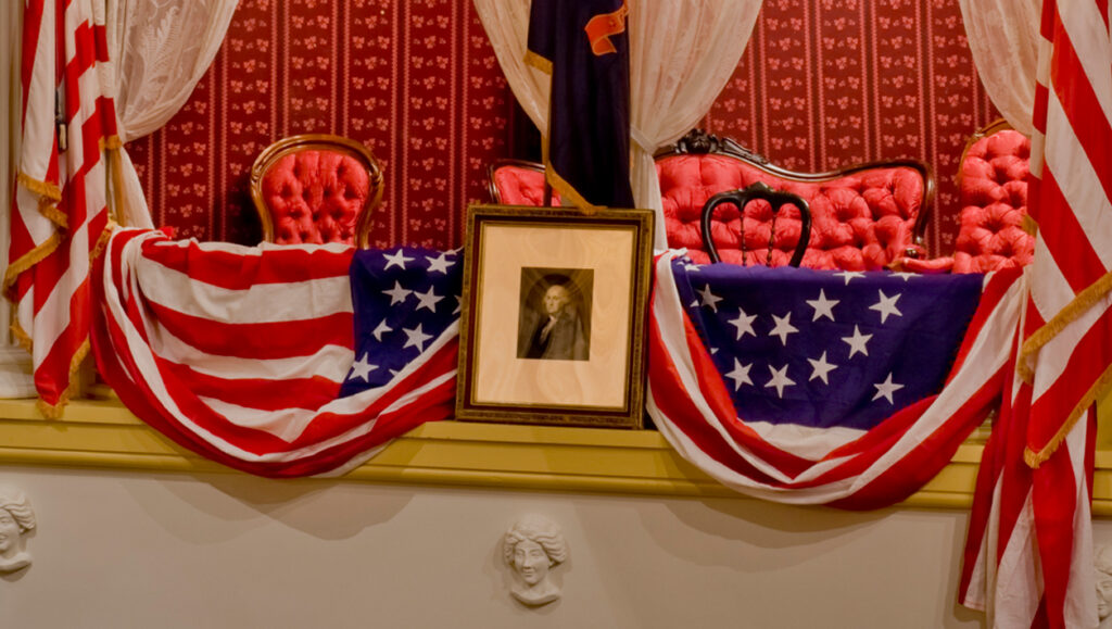 The President’s Box at Ford’s Theatre with American flags on either side, a picture of George Washington in the center and American flag bunting draped over the box. The interior of the box has red patterned wallpaper and red upholstered chairs and a couch. The box is framed by gold curtains with white lace curtains underneath them.