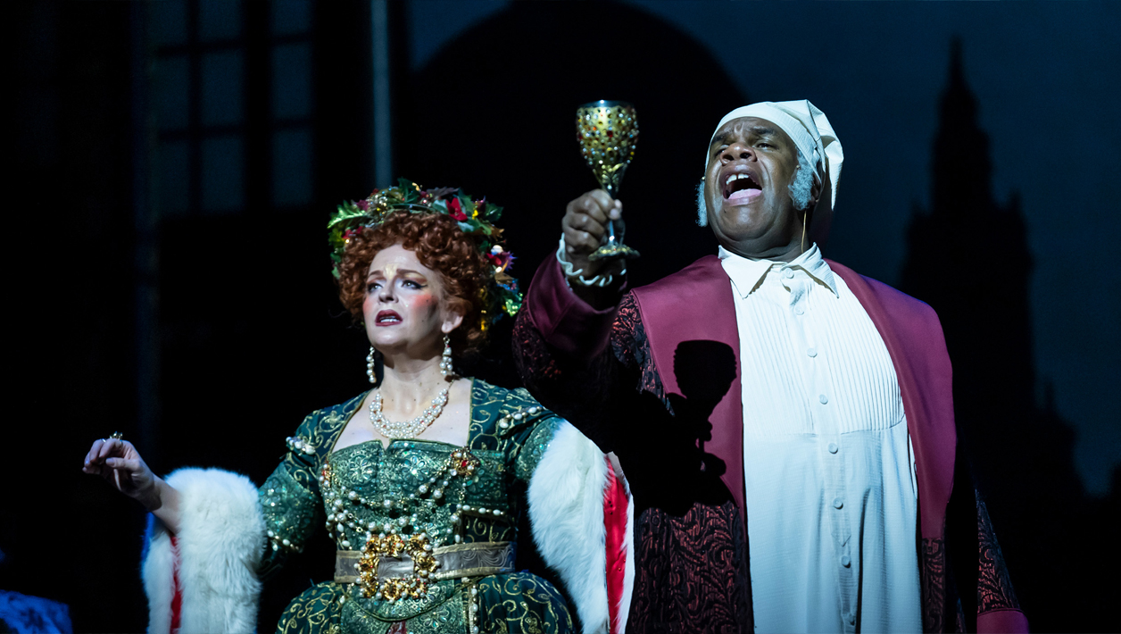 A Black man wearing a Victorian-era night cap, white nightgown and red house robe and suit holds up a goblet with fervor, as a spirit wearing a lavish Victorian-era green gown and wreath crown gazes with a serious expression.