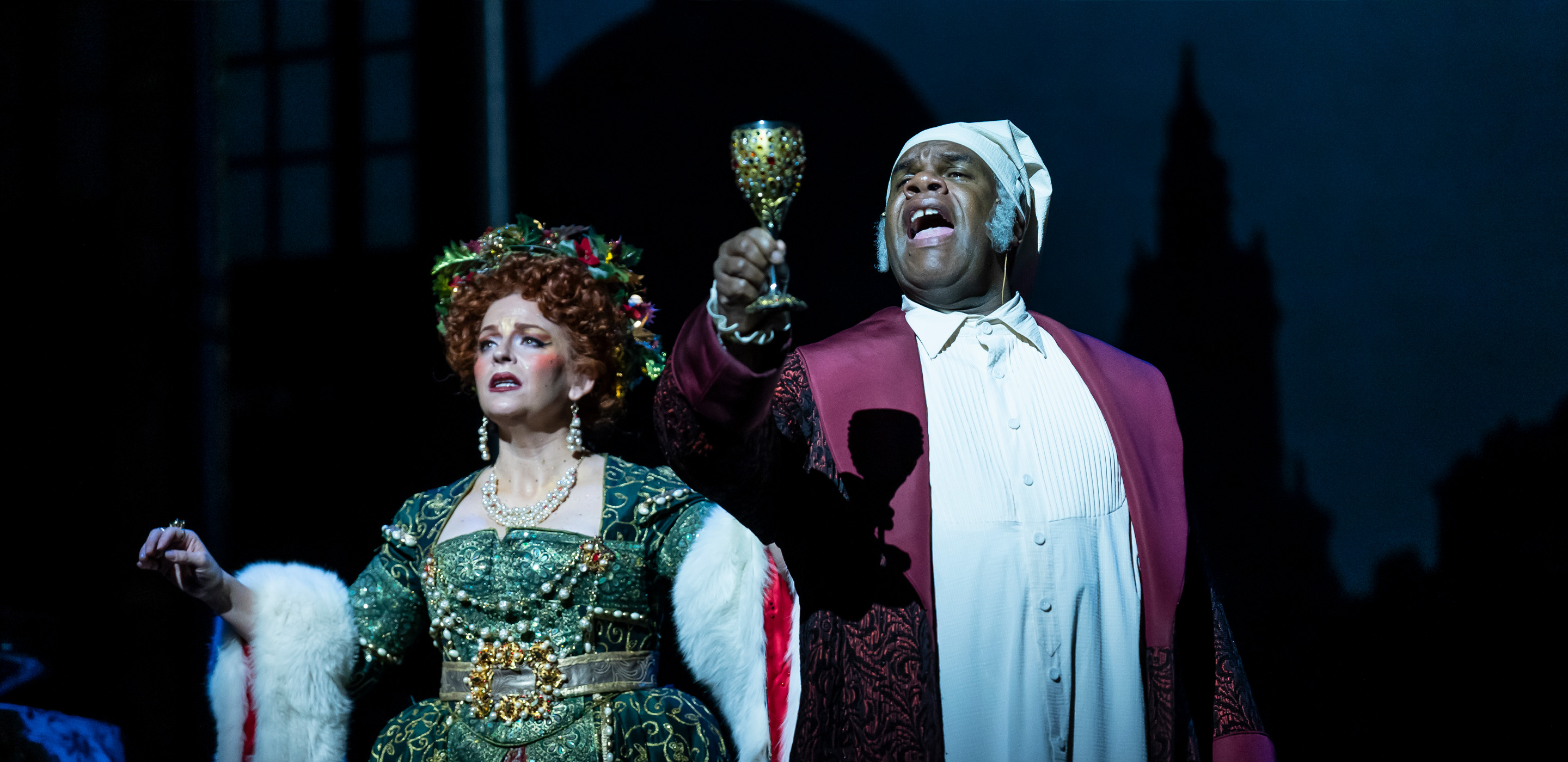 A Black man wearing a Victorian-era night cap, white nightgown and red house robe and suit holds up a goblet with fervor, as a spirit wearing a lavish Victorian-era green gown and wreath crown gazes with a serious expression.