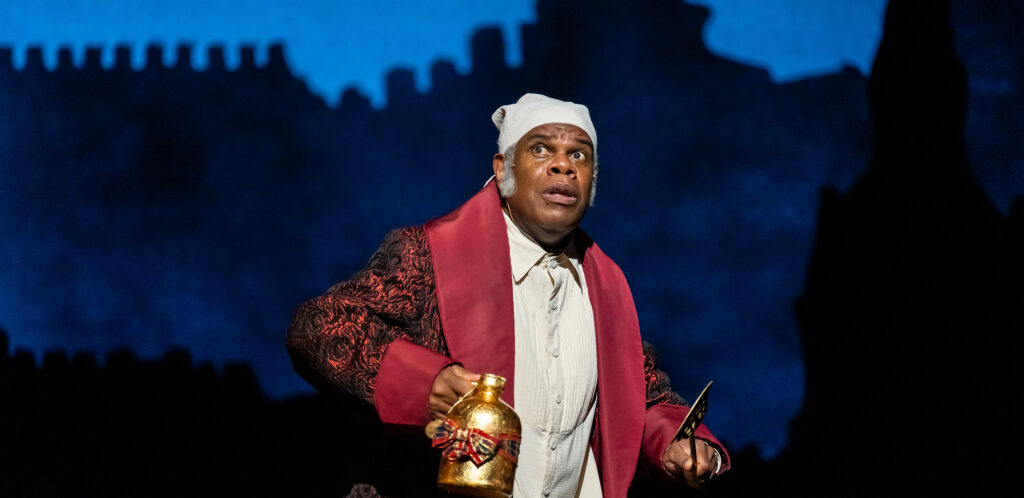 A Black man wearing a Victorian-era night cap, white nightgown and red house robe with fear on his face is holding a shiny brass pitcher.