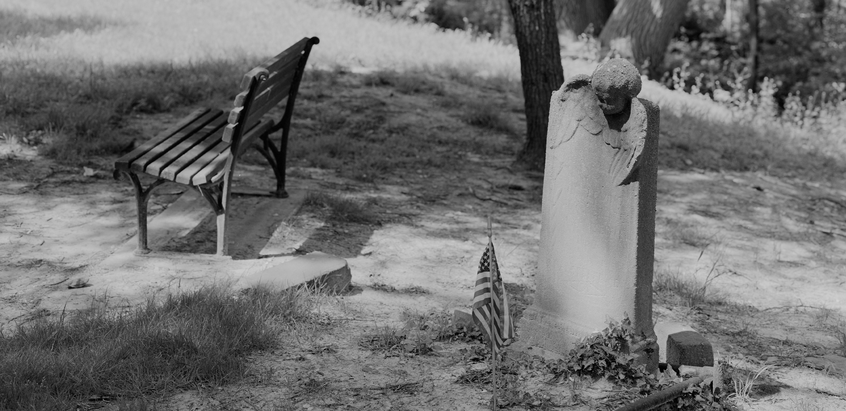 A black and white photograph of a grassy hill with a stone headstone topped by a cherub’s head and wings. A small American flag stands in the earth, decorating the grave. A park-bench is nearby on the left.