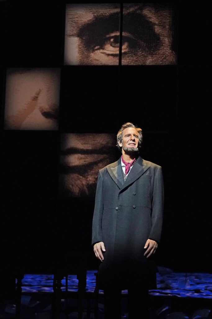 Abraham Lincoln, dressed in a black coat and red kerchief, stands in front of a partial picture of his face.