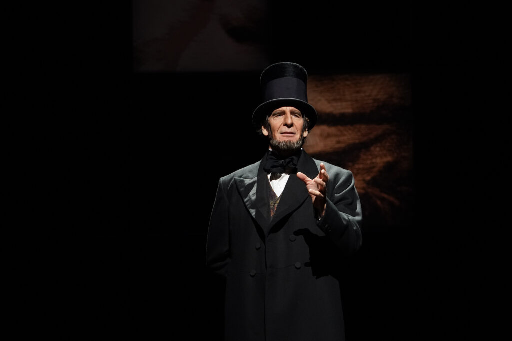 Abraham Lincoln, wearing a black coat, black tie and top hat, stands and speaks.