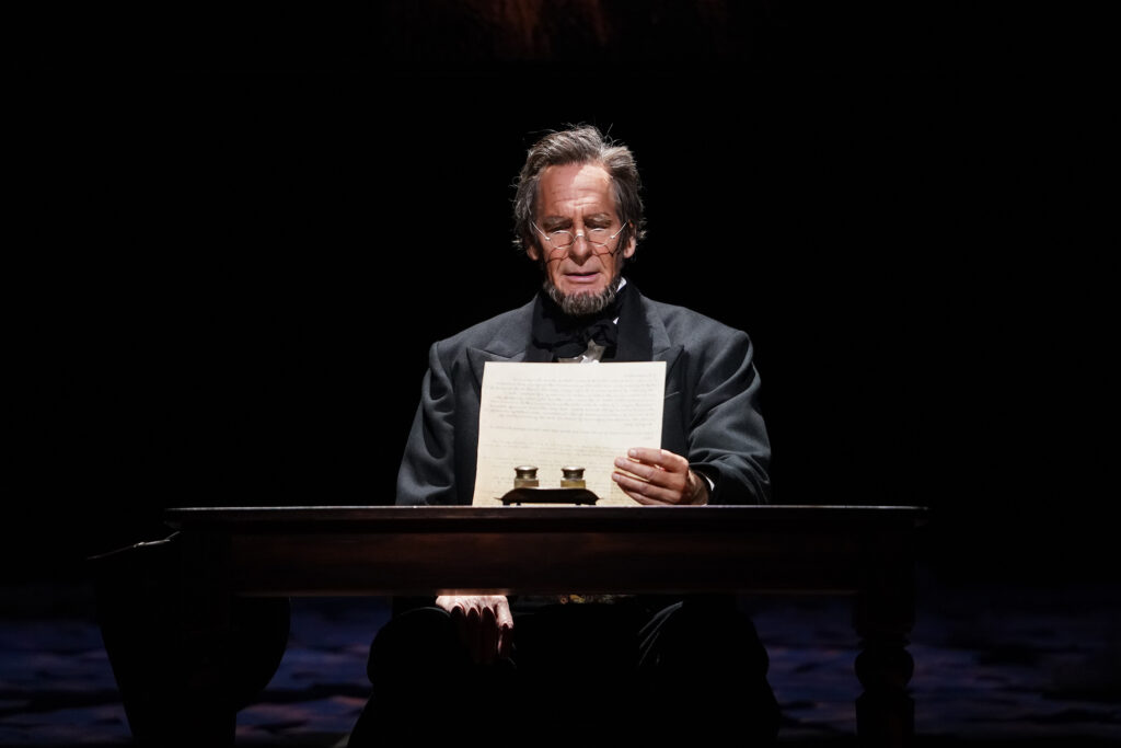 Abraham Lincoln, dressed in a black coat and tie, sits at a desk and reads from a piece of paper.