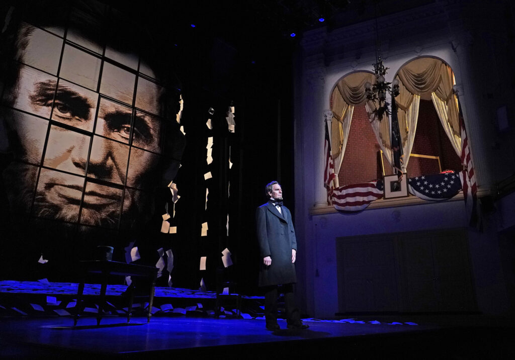 Abraham Lincoln stands on a stage with a large picture of his face to his left and a lit seating box to his right.