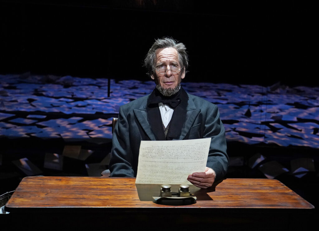 Abraham Lincoln, dressed in black coat and tie and wearing glasses, sits at a desk and holds a piece of paper while looking into the distance.