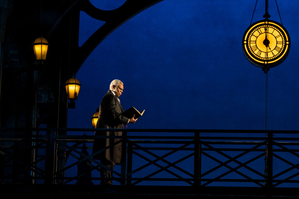 A Black man wearing a black coat looks at the pages of a book in his hand. He stands in the dark near lamp posts under a Victorian-era clock.