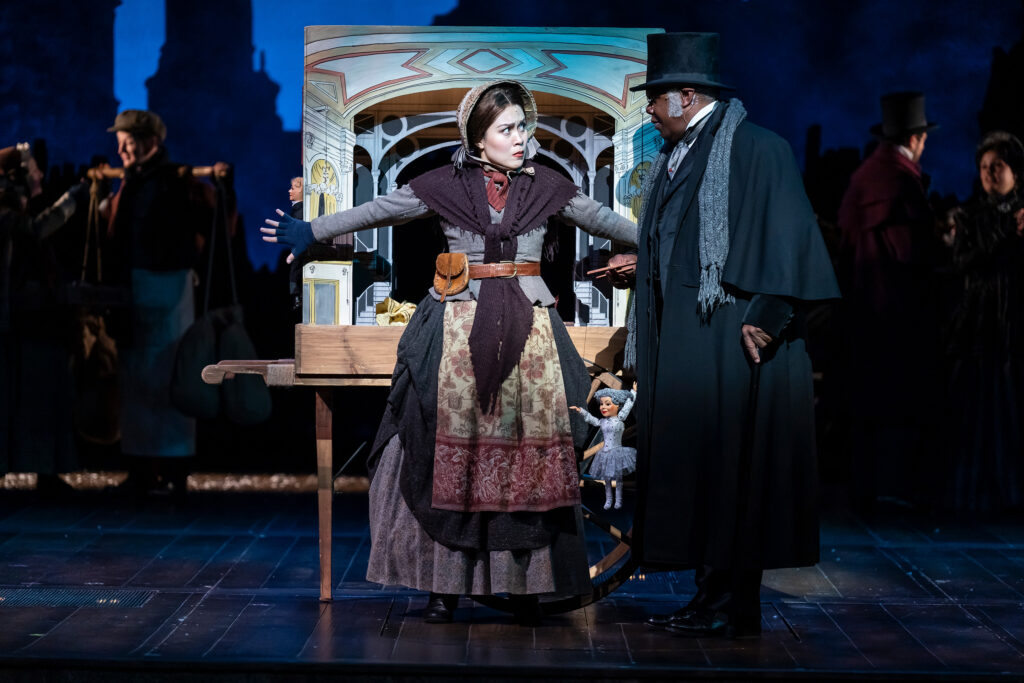 A determined vendor stands with arms outstretched to protect the Victorian-era doll cart behind her from a well-dressed serious-looking man in a black coat and top hat.