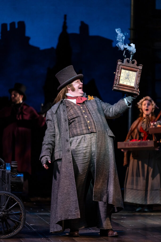 A smiling white man in a top hat and long grey jacket holds a Victorian-era square wooden clock with a line of smoke emerging from the top. E-News/Publications: Joe Mallon in A Christmas Carol (2024). Photo by Scott Suchman. Joe Mallon as Clock Vendor in the 2024 Ford’s Theatre production of “A Christmas Carol,” adapted by Michael Wilson, directed by Michael Baron. The production plays November 21-December 31, 2024, and features Scenic Design by Lee Savage, Costume Design by Alejo Vietti, Lighting Design by Rui Rita, Original Music and Sound Design by Josh Schmidt, Choreography by Shea Sullivan and Wig Design by Charles G. LaPointe. Photo by Scott Suchman. A smiling white man in a top hat and long grey jacket holds a Victorian-era square wooden clock with a line of smoke emerging from the top.