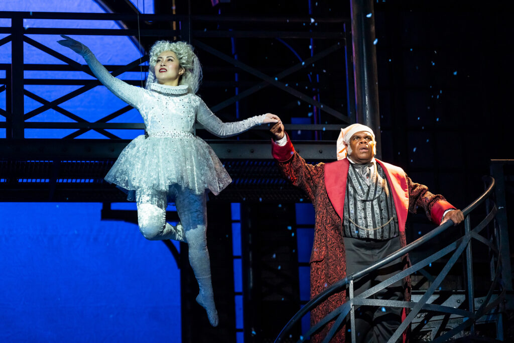 A Black man wearing a Victorian-era night cap and red house robe holds the hand of an Asian woman dressed as the Ghost of Christmas Past in all white who gracefully floats in the air.