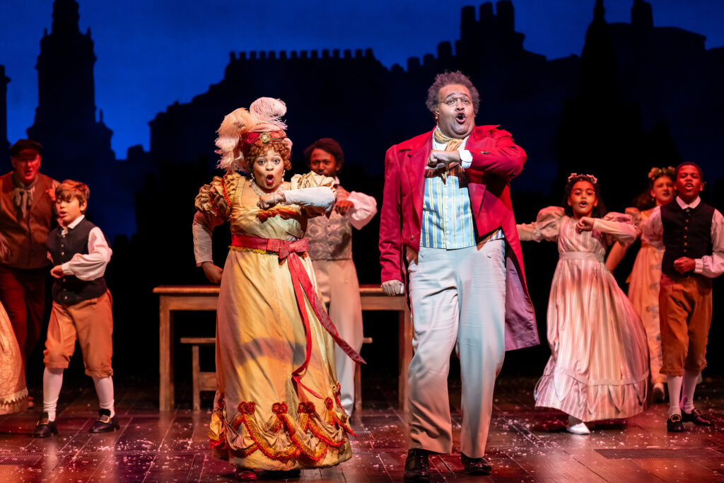 A woman and a man dressed in red and gold elegant Victorian-era party clothes dance at a holiday party.