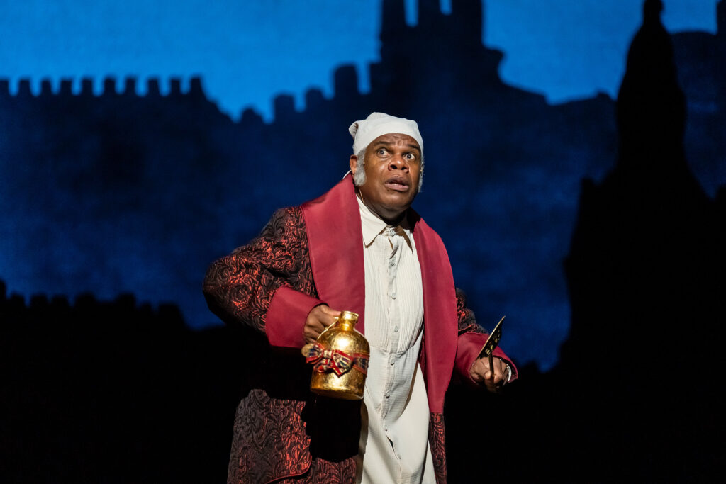 A Black man wearing a Victorian-era night cap, white nightgown and red house robe with fear on his face is holding a shiny brass pitcher.